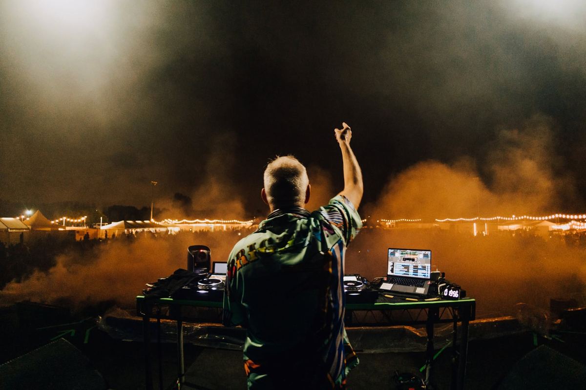 Fatboy Slim at the decks seen from behind, arm raised to a smoky festival field strung with lights